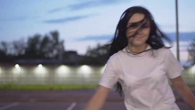 Young carefree woman riding to camera on rollers. Portrait of joyful Caucasian skater enjoying evening ride at dusk. Happy smiling woman skating on empty parking lot in city.