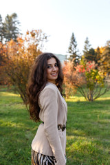 Beauty portrait of a cheerful brunette girl with long curly hairstyle in elegant fall outfit, walking in sunny day in the park.