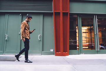 Young man in trendy clothes walking and using smartphone on stret