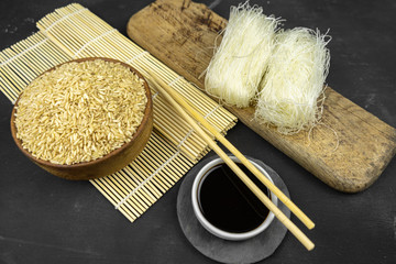 brown rice in a wooden bowl, asian kitchen background with bamboo mat, chopsticks, soy sauce, pickled ginger and glass rice noodles on black background