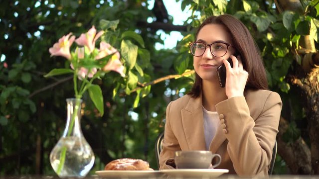 Young business woman sits in a cafe and speaks on the phone. Girl with coffee and a bun on the summer veranda.