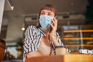 Pretty female in mask is looking away and talking on smartphone in cafe