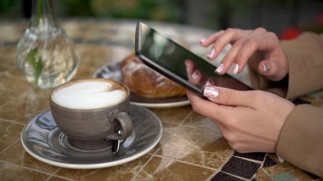 Young business woman sits in a cafe and writes text messages on a tablet close-up. Girl with coffee and a bun on the summer veranda.