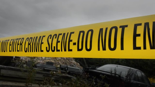 Police Crime Scene Cordon Tape Around Junkyard Car.  There is an overcast sky and light rainfall. Suggestive of the scene of a murder or hiding of a body.