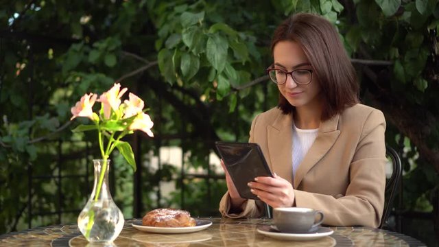 Young business woman sits in a cafe and writes text messages on a tablet. Girl with coffee and a bun on the summer veranda.