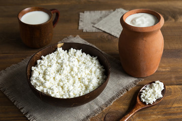 In wooden and clay dishes dairy products cottage cheese, milk, sour cream close up. The bowl stands on canvas napkin against a dark wooden background.