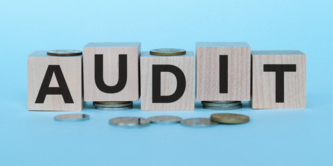 AUDIT word written on wooden blocks on stacks of coins on light blue backround. Selective focus. Top view.