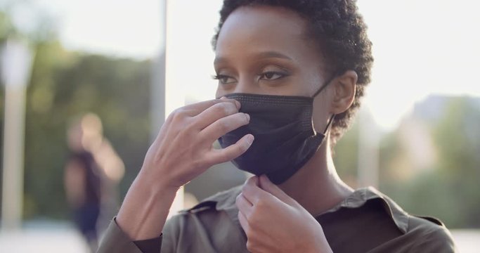 Portrait of isolated ethnic girl standing in street putting on black medical mask on female African face, protecting herself from covid virus, pandemic time, pollen allergy seasonal disease, close up