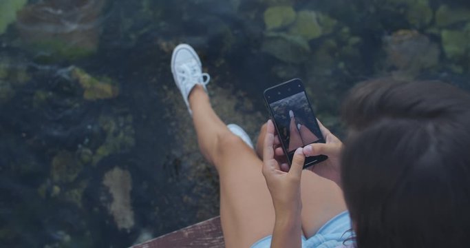 Happy woman sits on a pier against the background of the sea and takes pictures of her legs on the phone. Recreation and tourism concept. View from above. High quality 4k footage