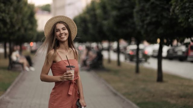 Beautiful young woman in a dress and hat walks in a city park, returns and smiles at the camera