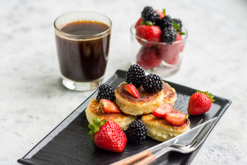 Cottage cheese pancakes with blackberries and strawberries on a dark plate with cutlery and a glass with coffee on a light background horizontal arrangement
