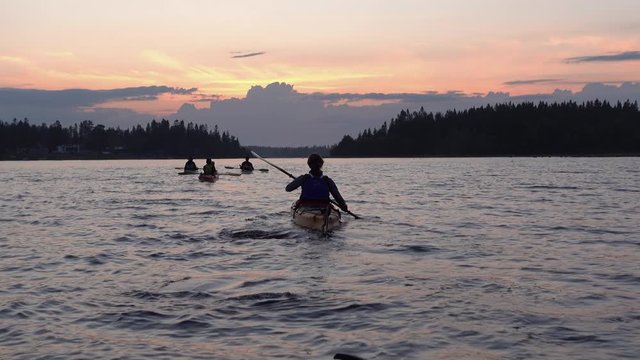 Young women in front of group of young people kayaking during light night in Swedish Lapland, calm water, summer forest. Healthy activity, midnight, Sun behind clouds