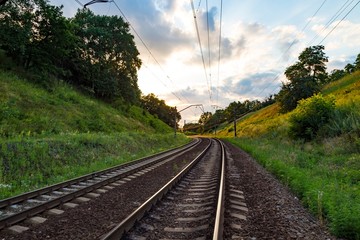 The railway line rails and electric poles and wires in the woodland in sunset time in summer.