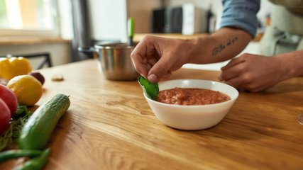 Close up of hands of man, cook serving traditional soup, using basil leaves for decorating the meal. Cooking at home, Italian cuisine concept