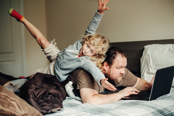 Work from home with kids children. Father working on laptop in bedroom with child daughter on his back. Funny candid family moments. New normal during coronavirus self-isolation quarantine. - 374003548