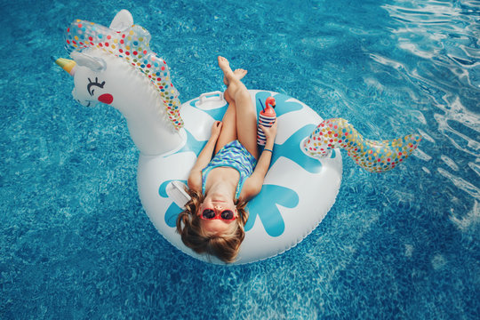 Cute adorable girl in sunglasses with drink lying on inflatable ring unicorn. Kid child enjoying having fun in swimming pool. Summer outdoors water activity for kids. View from above overhead.