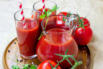 Fresh red tomato juice in a glass with a straw and jar with tomatoes on light wooden background.