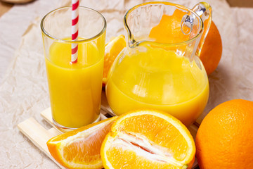 Fresh orange juice in a glass with a straw and jar with fruit pieces on light wooden background.