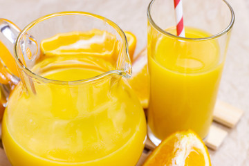 Fresh orange juice in a glass with a straw and jar with fruit pieces on light wooden background.