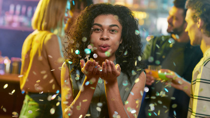 Attractive mixed race young celebrating woman blowing confetti to camera while posing in the night club. Friends chatting, having drinks at the bar counter in the background