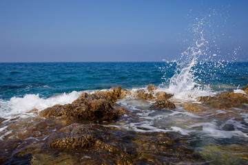 Wonderful views of the blue Mediterranean Sea. Sunny rocks, waves with foam and splashing water. The wave crashes into the rocks on the shore