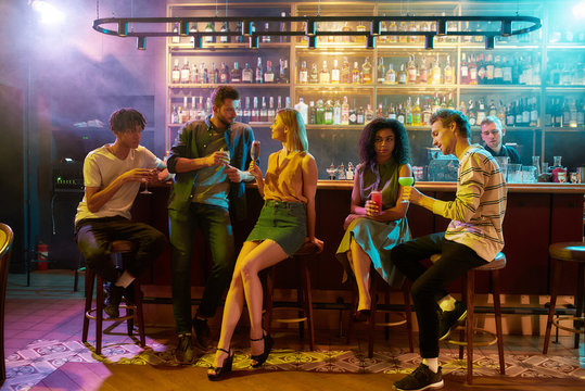 Gathering place. Full length shot of young men drinking cocktail at the bar counter and flirting with women they just met. Young adults having drinks while chatting at the night club