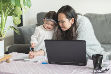 Asian woman working from home helps her daughter draw on a notepad while working on her laptop computer, sitting on a sofa in the living room of a house in Edinburgh, Scotland, UK, as both smile