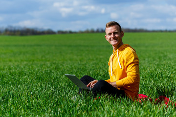 Naklejka na ściany i meble Young businessman sitting on green grass and using laptop computer. Handsome man working with computer in park at sunny summer day. Outdoors nature journey and relaxation. Freelance work concept