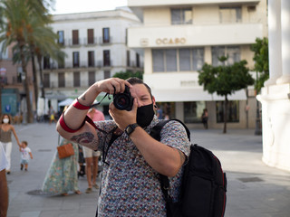 Fotógrafo hombre joven con tatuajes tomando una foto en una calle con edificios de oficinas vistiendo mascarilla durante la nueva normalidad