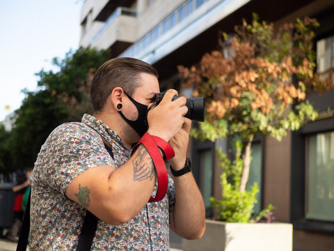 Fotógrafo hombre joven con tatuajes tomando una foto en una calle con edificios de oficinas vistiendo mascarilla durante la nueva normalidad