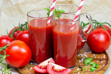 Fresh red tomato juice in a glass with a straw and jar with tomatoes on light wooden background.