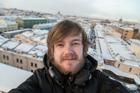 Portrait of a funny fair-haired boy with disheveled hair and a beard on the background of the snow-capped rooftops of St. Petersburg