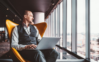 A handsome cheerful caucasian man entrepreneur is sitting with a laptop on his knees on an orange armchair next to the window indoors of a luxurious interior of a modern business office skyscraper