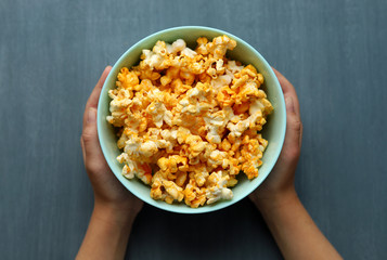 A blue dish full of popcorn in the hands of a Caucasian man on a gray background. Selective focus.