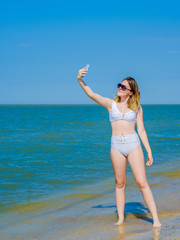 Beautiful young girl on the sea beach near the water posing and taking a selfie on a smartphone. Girl in a white swimsuit and sunglasses. Summer sunny day. Sea vacation concept. Copy space.