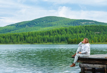 Fototapeta na wymiar A young woman sits on a wooden pier at a high mountain lake against the backdrop of mountains in the Altai Republic in Russia