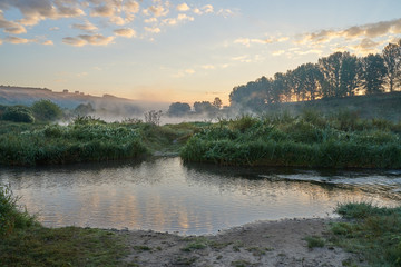 Summer time. Morning dawn over the river in a hazy, thoughtful haze. Beautiful view of the forest and river covered with fog early in the morning.
