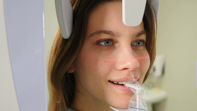 Young smiling female patient undergoing panoramic X-ray investigation of oral cavity using an orthopantomographic machine. Laser lines on face. Ct scanner in modern dental clinic. 4 k video