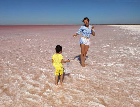 The coast of the salty Pink Lake in the Crimea. A young woman in shert and a little boy in yellow clothes carries a bottle of water to his mother against the background of the lake. Mother and son. 