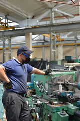 Fototapeta na wymiar Working turner wearing a protective antiviral mask on a lathe in a workshop. The machine operator at the workplace controls the equipment.