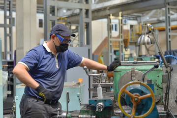 Working turner wearing a protective antiviral mask on a lathe in a workshop. The machine operator at the workplace controls the equipment.