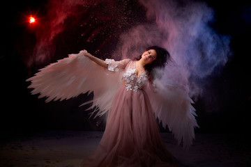 Naklejka premium Girl with white wings posing in dark black studo during photoshoot with flour or dust and light. White angel in dark space