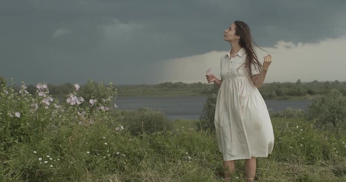 Elegant girl stroking long hair on summer wind rainy country landscape background. Young woman in white dress standing alone on nature outdoors slow motion copy space. Change freedom concept
