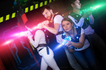 Modern young people playing laser tag on dark labyrinth in bright beams of laser pistols