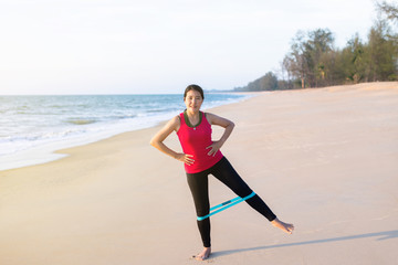 Happy asian woman using resistance band exercises for stronger legs at the beach
