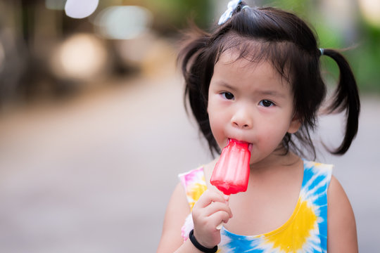 Happy Adorable Little Asian Child Eating Red Popsicle In The Summer Or Spring Time. Head Shot Of Funny Girl Enjoy Eating Jelly Ice Cream. At The Street. Cute Kid Aged 3 Years Old.