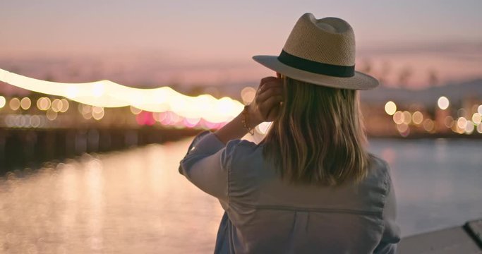 Slow motion of woman enjoying view on promenade lighting. Back view of stylish young female traveler in hat enjoying evening on the pier with night city view. Female traveler exploring USA coast