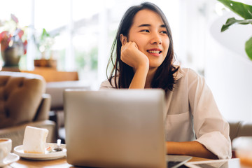 Portrait of smiling happy beautiful asian woman relaxing using laptop computer while sitting on sofa.Young hipster girl freelancer working and thinking with new ideas in cafe and restaurant