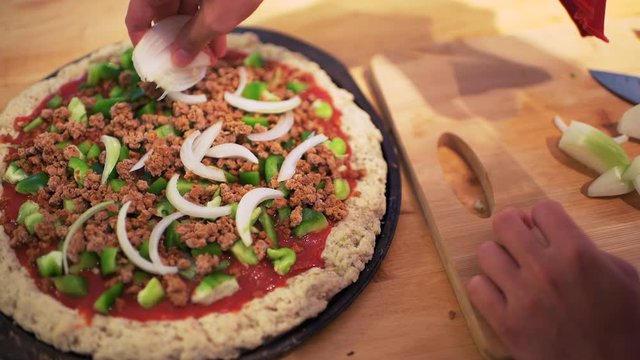 Flat top view down of raw pizza dough crust before baking made with gluten free flour and woman hands adding chopped green bell peppers and onions on red tomato sauce 