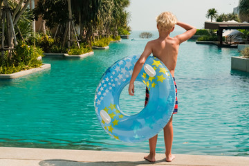 Happy little boy standing near the swimming pool with a blue and yellow pool float in his hands. Copy space banner.